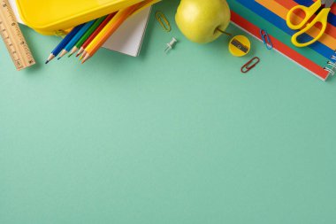 Okul malzemeleri, yeşil masa arkasına yayılmış kalem, elma ve makas gibi.