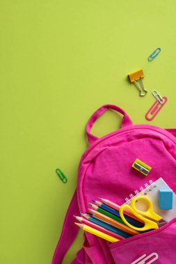 Okul malzemeleriyle dolu canlı, dikey pembe bir sırt çantası yeşil bir yüzeye oturmuş.
