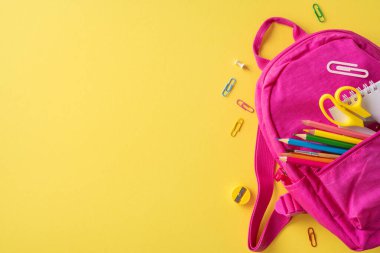 Parlak, pembe bir sırt çantası ve parlak sarı bir zemin üzerinde renkli kırtasiye malzemeleri.