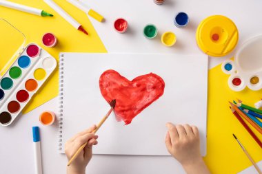 Bir çocuk rengarenk sanat malzemeleriyle çevrili kırmızı bir kalp yapar.