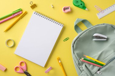Okul malzemelerinin bir koleksiyonu, içinde bir defter, sırt çantası ve renkli kırtasiye, neşeli sarı bir yüzey üzerinde sergileniyor.