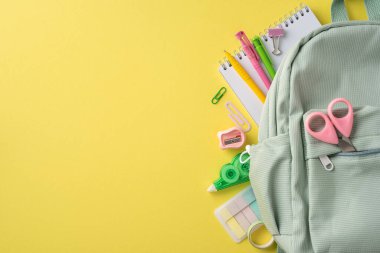 Canlı sarı bir yüzey üzerinde çeşitli renkli okul malzemeleriyle dolu yeşil bir sırt çantası.