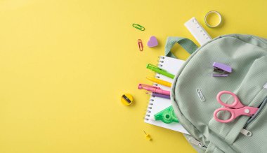 Çeşitli renkli okul malzemeleri ve parlak sarı bir yüzeye yerleştirilmiş yeşil bir sırt çantası.