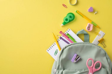 Kırtasiye malzemeleri, kalemler, defterler ve bir sırt çantası da dahil olmak üzere okul malzemeleri parlak sarı bir arkaplanda yaratıcı bir şekilde düzenlenmiştir.