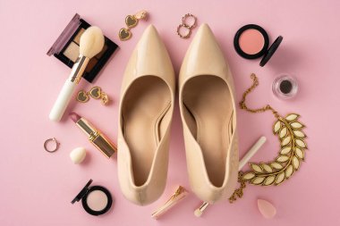 Pembe bir yüzeyde çeşitli kozmetik ürünler ve aksesuarlarla çevrili bej renkli kadın ayakkabıları.