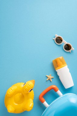 Mavi arka planda bir valiz, güneş kremi, güneş gözlüğü, denizyıldızı ve şişme ördek de dahil olmak üzere yaz seyahati aksesuarları içeren düz bir bileşim yatıyordu.