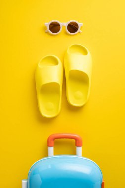 Sarı sandaletler, güneş gözlükleri ve rahatlama ve tatil hazırlıklarını temsil eden mavi bir bavul içeren parlak, dikey yaz seyahati öğeleri