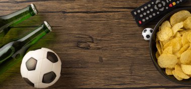 Futbol topu, bira şişeleri, uzaktan kumanda ve kırsal bir ahşap masada bir kase cips içeren bir düzenleme, spor müsabakası izleme veya boş vakitlerin keyfini çıkarma havasını iletiyor.
