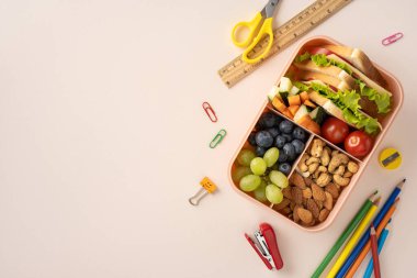 Parlak bir yüzeyde okul malzemelerinin yanında sandviç, meyve ve fındık içeren düzenli bir beslenme çantası.