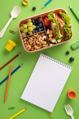 Okul malzemeleriyle çevrili meyve, fındık, sebze ve sandviçin bulunduğu dikey sağlıklı bir beslenme çantası. Eğitimi ve besleyici yeme alışkanlıklarını vurgulayan yeşil arka planda.