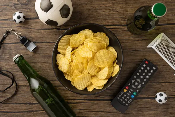 Bira şişeleri, uzaktan kumanda, ıslık ve futbol temalı eşyalar ile çevrili bir masada patates cipsi.
