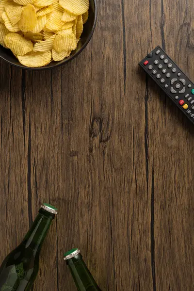 Bira şişeleri, patates cipsleri ve uzaktan kumandalı bir ev rahatlama konseptini tasvir eden dikey bir sahne.