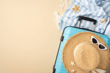 Mavi bavul, hasır şapka ve tatil eşyalarının yer aldığı bir tatil düzeninin üst görüntüsü