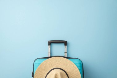 Turkuaz bavulun üzerinde açık mavi bir arka planda duran hasır bir şapka var.