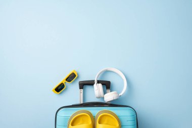 Mavi bir bavul, kulaklık, terlik ve mavi arka planda duran güneş gözlüğü de dahil olmak üzere renkli yaz tatili esasları.