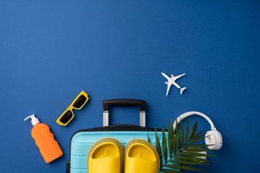 Bir bavul, kulaklık, güneş gözlüğü ve mavi arka planda terliklerle tatil için gerekli şeyler.