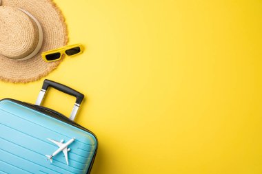 Hasır şapka, bavul, güneş gözlüğü ve oyuncak uçak, somutlaştırıcı seyahat ve yaz tatili titreşimleri içeren parlak sarı bir arkaplan.