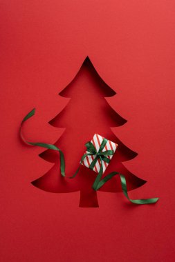 Çizgili hediye kutusu olan bir Noel ağacı kesiminin olduğu sanatsal dikey kırmızı bayram kartı. Zarif kompozisyon şenlik ruhunu ve mevsimsel neşeyi çağrıştırıyor.
