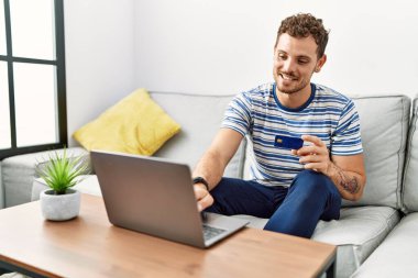 Young hispanic man using laptop holding credit card at home