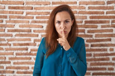 Brunette woman standing over bricks wall asking to be quiet with finger on lips. silence and secret concept. 