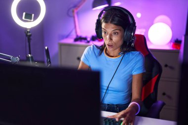 Brunette young woman playing video games scared and amazed with open mouth for surprise, disbelief face 