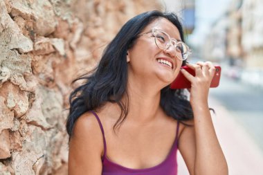 Young chinese woman smiling confident listening audio message by the smartphone at street
