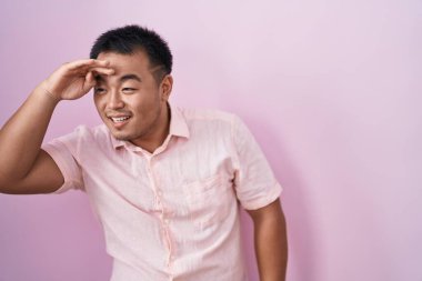 Chinese young man standing over pink background very happy and smiling looking far away with hand over head. searching concept. 