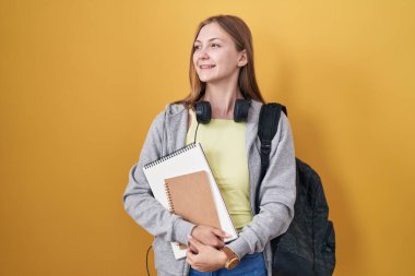Young caucasian woman wearing student backpack and holding books looking away to side with smile on face, natural expression. laughing confident. 