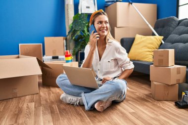 Young hispanic woman using laptop and talking on smartphone at new home