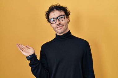 Hispanic man standing over yellow background smiling cheerful presenting and pointing with palm of hand looking at the camera. 