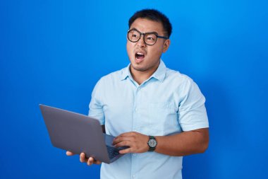 Chinese young man using computer laptop angry and mad screaming frustrated and furious, shouting with anger. rage and aggressive concept. 