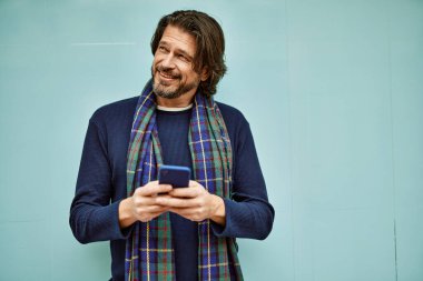 Orta yaşlı yakışıklı adam duvara dayanmış akıllı telefon kullanıyor.