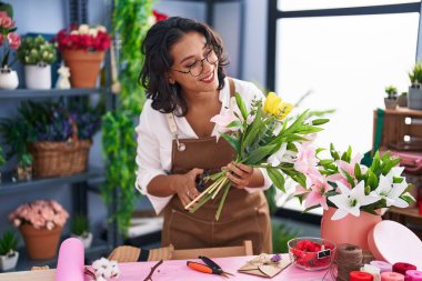 Young beautiful hispanic woman florist make bouquet of flowers at flower shop