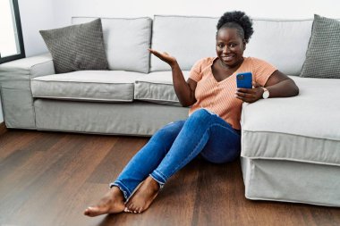 Genç Afrikalı bir kadın akıllı telefon kullanıyor. Evinde oturmuş gülümsüyor ve avucunun içi kameraya bakıyor.. 