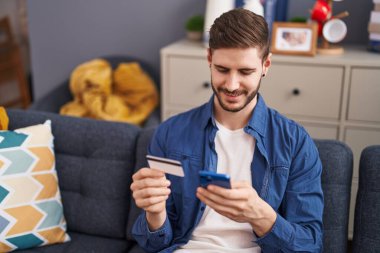 Beyaz bir adam akıllı telefon ve kredi kartı kullanıyor. Evdeki kanepede oturuyor.