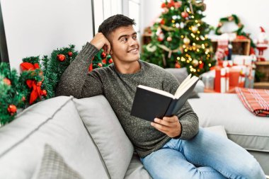 Genç İspanyol adam kitap okuyor. Evdeki Noel ağacının yanında, koltukta oturuyor.