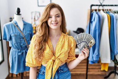 Young caucasian woman at retail boutique holding dollars banknotes looking positive and happy standing and smiling with a confident smile showing teeth 