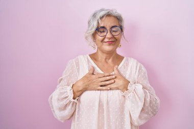 Middle age woman with grey hair standing over pink background smiling with hands on chest with closed eyes and grateful gesture on face. health concept. 