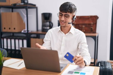 Young hispanic man working using computer laptop holding credit card smiling cheerful offering palm hand giving assistance and acceptance. 