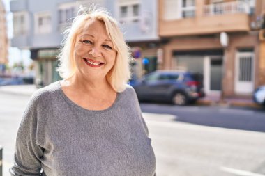 Orta yaşlı sarışın kadın sokakta kendinden emin bir şekilde gülümsüyor.