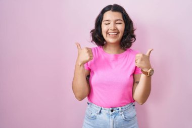 Young hispanic woman standing over pink background success sign doing positive gesture with hand, thumbs up smiling and happy. cheerful expression and winner gesture. 