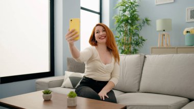 Genç kızıl saçlı kadın akıllı telefonuyla özçekim yapıyor. Evdeki kanepede oturuyor.