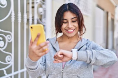 Young hispanic woman wearing sportswear looking stopwatch using smartphone at street