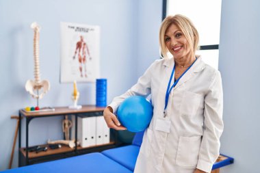 Middle age blonde woman wearing physiotherapist uniform holding ball at physiotherapy clinic