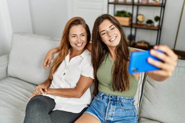 Mother and daughter smiling confident and hugging each other making selfie by the smartphone at home