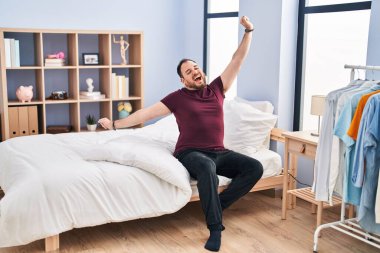 Young hispanic man waking up sitting on bed at bedroom