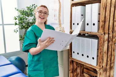 Fsiolog üniforması giyen genç kızıl saçlı kadın klinikte klasör tutuyor.