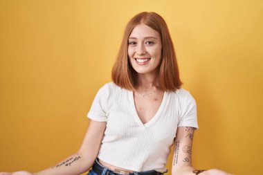 Young redhead woman standing over yellow background smiling cheerful with open arms as friendly welcome, positive and confident greetings 