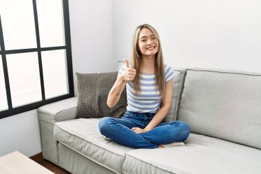 Asian young woman sitting on the sofa at home doing happy thumbs up gesture with hand. approving expression looking at the camera showing success. 