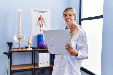 Fizyoterapi kliniğinde fizyoterpist üniforması giyen genç sarışın kadın tıbbi rapor okuyor.